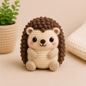 May include: A small, round, handmade crochet hedgehog toy featuring a textured dark brown back and a cream-colored face, belly, and limbs. The figure has two black plastic eyes, a small brown nose, a stitched mouth, and two pink blush spots.