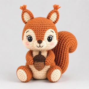 May include: A crocheted squirrel toy featuring a rust-orange body with cream-colored accents on the face, chest, and feet. The squirrel has large black eyes, pink cheeks, tufted ears, and holds a small brown acorn in its front paws.