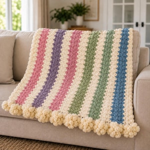 May include: A crochet blanket featuring vertical stripes in cream, pink, purple, green, and blue. The blanket has a textured stitch pattern and is finished with a decorative border of cream-colored pom-poms along the bottom edge.