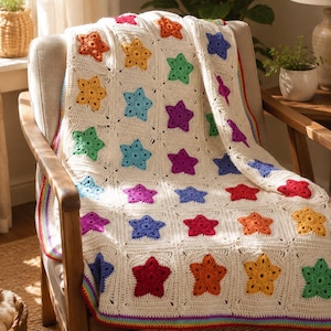 Può includere: Una coperta all'uncinetto color crema caratterizzata da un motivo ripetuto di stelle multicolori nelle tonalità del rosso, arancione, giallo, verde, blu e viola. La coperta è rifinita con un vivace bordo a righe arcobaleno lungo i lati.