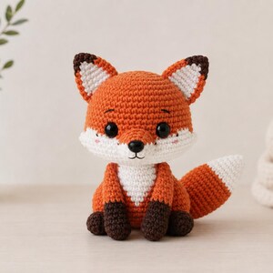 Puede incluir: Un peluche de zorro tejido a ganchillo con cuerpo naranja brillante, pecho y cara blancos, patas de color marrón oscuro y orejas puntiagudas con centros blancos. El zorro tiene grandes ojos negros de seguridad, una pequeña nariz negra y una cola tupida con la punta blanca.