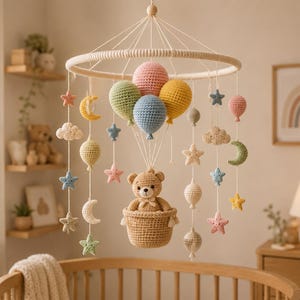 May include: A crochet baby mobile featuring a central tan teddy bear sitting in a basket suspended by strings from a circular frame. The mobile includes various crocheted elements such as colorful balloons, stars, moons, and clouds in pastel tones.