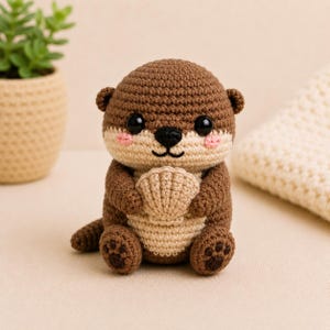 Puede incluir: Un peluche de nutria tejido a ganchillo en color marrón y beige con cabeza redonda, ojos de plástico negro, una pequeña nariz negra y mejillas bordadas en rosa. La nutria sostiene una concha tejida en beige y tiene almohadillas marrones.