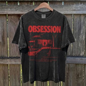 May include: A charcoal grey short-sleeve t-shirt with a vintage wash finish. The front features the word OBSESSION in large red letters above a graphic of a house and a car. Small red text is printed below the graphic on the chest area.