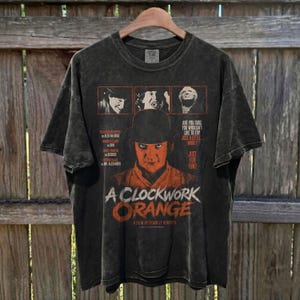 May include: A charcoal gray acid-wash t-shirt featuring a graphic print from the film A Clockwork Orange. The design includes three small character portraits at the top, a central portrait of a person in a bowler hat, and the text A Clockwork Orange.