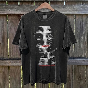 May include: Dark gray t-shirt with a graphic of multiple eyes and the word "OBSESSION" in red. The text "be careful what you wish for" is below the graphic. The shirt is hanging on a wooden hanger.