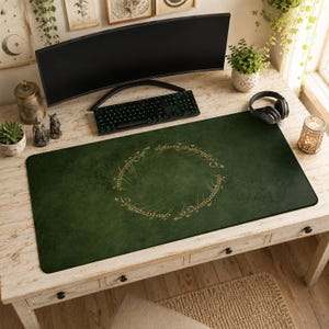 Può includere: Un tappetino verde per scrivania con un'iscrizione dorata a forma di cerchio. Il tappetino è rettangolare e poggia su una scrivania in legno chiaro. Sono visibili anche una tastiera e un monitor neri.
