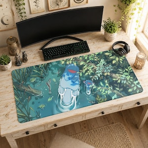 May include: A rectangular desk mat featuring a digital illustration of a blue, cartoonish axolotl-like creature swimming in a pond. The scene includes green aquatic plants, tree branches, and a smaller creature of the same type in the background.