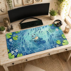 Op de afbeelding: Een rechthoekige bureaumat met een lichtblauw wateroppervlak waarop een lichtblauw schildpadfiguur zwemt tussen kleine blauwe vissen, groene waterleliebladeren en witte en blauwe waterleliebloemen, wat een serene aquatische scène vormt.