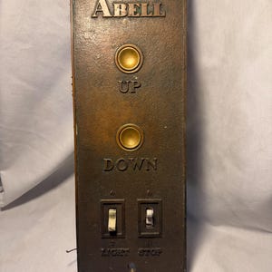 May include: A rectangular bronze-toned metal elevator control panel featuring the embossed text ABELL at the top. It includes two circular gold-colored push buttons labeled UP and DOWN, and two toggle switches below labeled LIGHT and STOP.