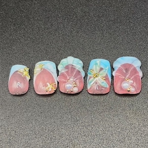 May include: A set of ten press-on nails featuring a pink base with light blue tips. The nails are decorated with three-dimensional white flower petals, small white pearls, and tiny gold metallic accents arranged in various patterns.