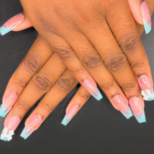 May include: Close-up of hands displaying long, square acrylic nails. The nails have a French manicure design with a light pink base and baby blue tips. Some nails are adorned with white flower accents. The background is a plain black.