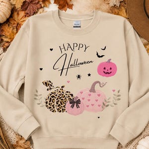 Könnte beinhalten: Ein beiges Langarm-Sweatshirt mit einem Grafikdruck und dem Text Happy Halloween. Das Design zeigt einen rosa Kürbis, einen Kürbis mit Leopardenmuster, einen rosa Kürbis mit Herzchen, eine kleine Spinne, Fledermäuse und kleine Herzen.
