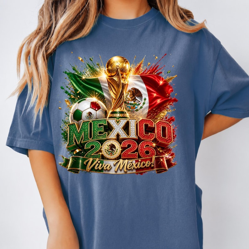 May include: A blue t-shirt featuring a graphic of the Mexico 2026 World Cup trophy, a soccer ball, and the Mexican flag. The text reads MEXICO 2026 and Viva M&eacute;xico! with a central emblem. The design includes colorful paint splatters and gold accents.