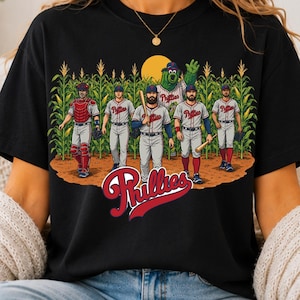 May include: A black t-shirt featuring a graphic of five baseball players and a green mascot standing in a cornfield. The players wear white uniforms with red lettering. The word Phillies is printed in large red script at the bottom of the design.
