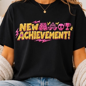 May include: A black short-sleeved t-shirt featuring the gold glitter text NEW ACHIEVEMENT! with pink glitter accents including lightning bolts, shorts, a tiara, sunglasses, a skull, and a trophy. The design is centered on the front of the shirt.