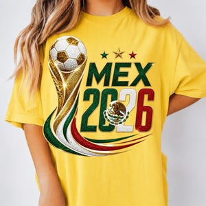 May include: A bright yellow t-shirt featuring a graphic of a soccer ball atop a gold trophy. The text reads MEX 2026, with the numbers colored in green, white, and red. The Mexican coat of arms is centered within the zero, and three stars appear above.