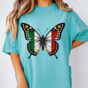 May include: A turquoise short-sleeve t-shirt featuring a large central graphic of a butterfly. The butterfly wings are colored with the green, white, and red vertical stripes of the Italian flag, accented with black outlines and small floral details.