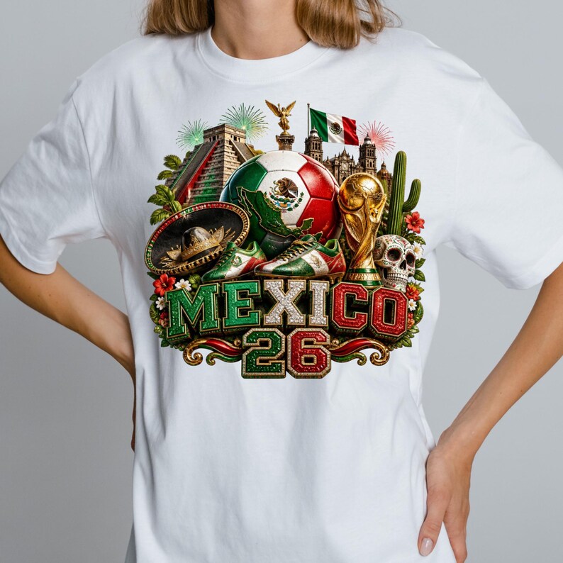May include: A white t-shirt featuring a colorful graphic print of a soccer ball, a trophy, a sombrero, a skull, a pyramid, and the Mexican flag. The text MEXICO 26 is displayed in large, textured, green, white, and red block letters at the bottom.