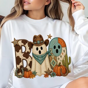 Puede incluir: Una sudadera blanca de cuello redondo con un estampado gráfico de la palabra BOO. La letra B tiene un patrón de estampado de vaca marrón, el fantasma lleva un sombrero de vaquero marrón y un pañuelo azul verdoso, y la O presenta un patrón naranja y azul verdoso con un cactus.