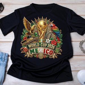 May include: A black short-sleeve t-shirt featuring a graphic print of the World Cup 2026 Mexico trophy, a golden eagle, a colorful parrot, a sombrero, a pyramid, and tropical flowers. The text reads World Cup 2026 Mexico in a decorative banner.