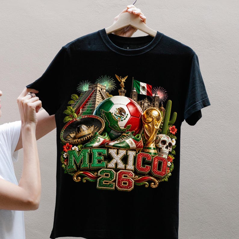 May include: A black t-shirt featuring a graphic print of a soccer ball with the Mexican flag, a gold trophy, a sombrero, a skull, a cactus, a pyramid, and the text MEXICO 26. The design includes fireworks and floral elements in red, white, and green.