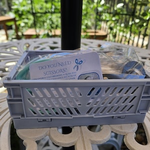May include: A gray plastic crate filled with various items, including a card that reads "Do You Need Scissors?" and a roll of blue tape. The crate sits on a white, ornate metal table, with greenery visible in the background.