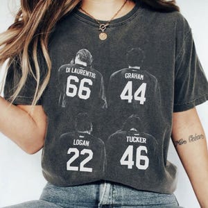 May include: Dark gray t-shirt featuring four jersey backs with names and numbers: Di Laurentis 66, Graham 44, Logan 22, and Tucker 46. The shirt has a vintage, worn look.
