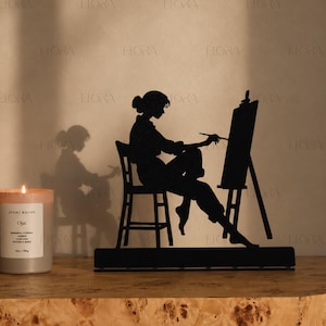Könnte beinhalten: Schwarze Metallsilhouette einer Person, die an einer Staffelei malt. Die Silhouette sitzt auf einem Stuhl, mit einer Kerze und Text auf der linken Seite. Die Kerze befindet sich in einem Glas. Der Text auf der Kerze lautet "Ojai".