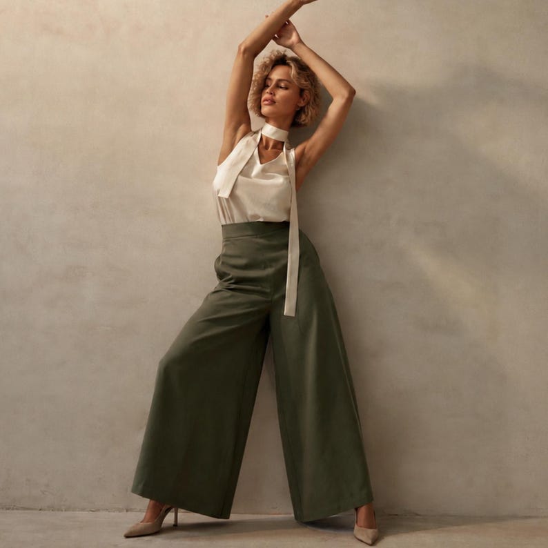 K&ouml;nnte beinhalten: Ein Damenoutfit mit einem wei&szlig;en Neckholder-Top mit langer, flie&szlig;ender Krawatte und einer olivgr&uuml;nen Hose mit weitem Bein. Das Outfit wird mit beigen Abs&auml;tzen kombiniert. Das Model lehnt an einer neutral get&ouml;nten Wand.