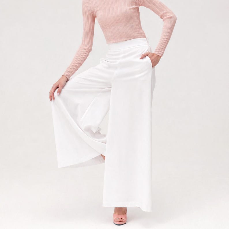 May include: A person wearing a light pink long-sleeved top and white wide-leg trousers. The trousers have pockets and a flowing design. The person is also wearing pink heels.