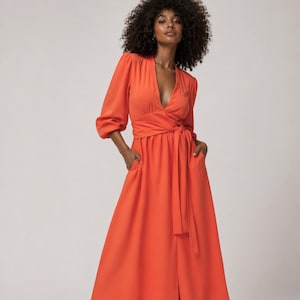May include: A vibrant orange wrap dress with a V-neckline and three-quarter sleeves. The dress features a waist tie and pockets. The long skirt has a front slit. The dress is made of a flowing fabric.