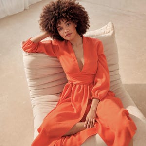 May include: An orange-red dress with long sleeves and a deep V-neck. The dress features a wrap-around waist and a flowing skirt. The model is seated on a beige, cushioned chair. The dress is made of a lightweight fabric.