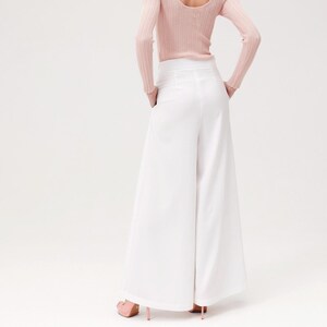 May include: A person wearing a pale pink long-sleeved top and white wide-leg trousers. The trousers have a high waist and a flowing shape. The person is wearing pink heels.