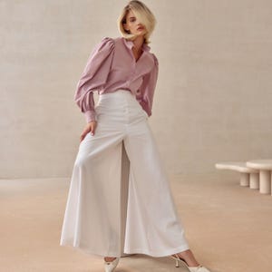 May include: A light mauve shirt with long, puffed sleeves and white wide-leg trousers. The trousers have a high waist and a split front. The model is wearing white heels.