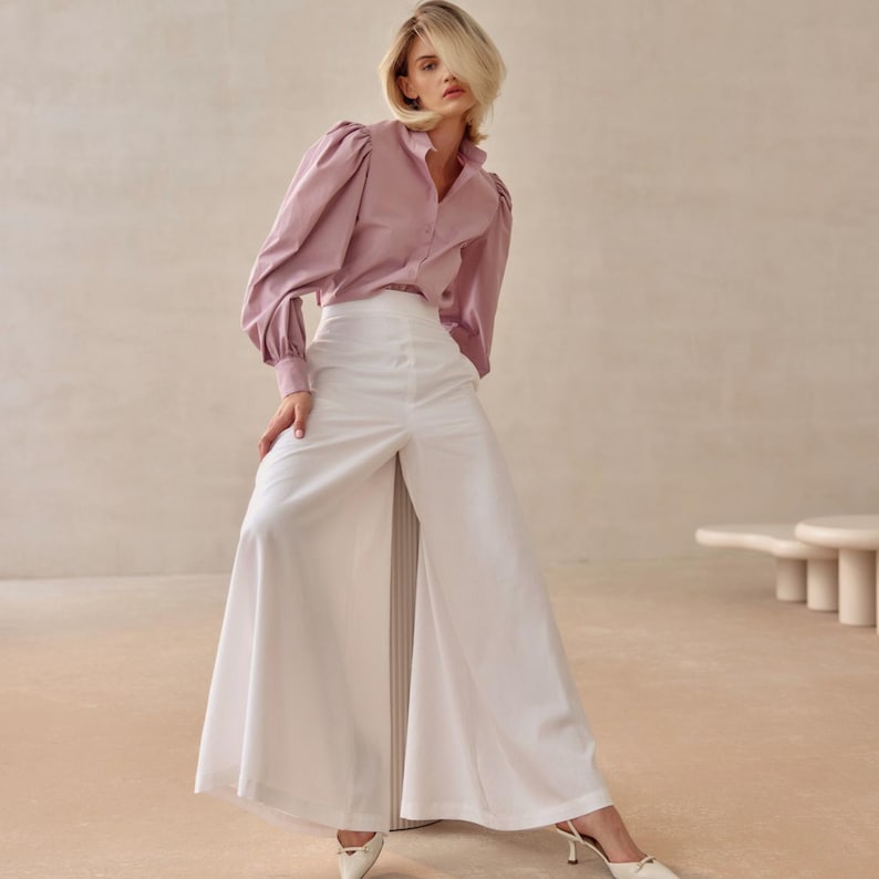 May include: A light mauve shirt with long, puffed sleeves and white wide-leg trousers. The trousers have a high waist and a split front. The model is wearing white heels.