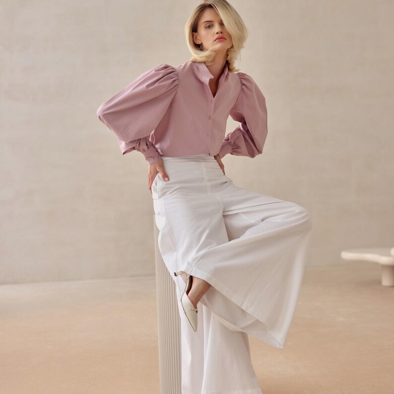 May include: A pale pink blouse with large, puffed sleeves and a button-down design. The blouse is worn with wide-legged white trousers and white shoes. The outfit is set against a neutral backdrop, creating a soft, elegant look. The blouse has a high collar.