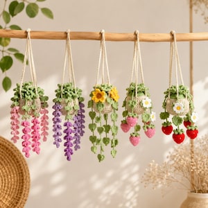 May include: Five crocheted hanging plant decorations suspended from a wooden branch. Each features a beige pot with green foliage and distinct dangling elements: pink flowers, purple wisteria, green heart-shaped leaves, pink strawberries, and red strawberries.