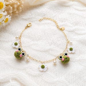Puede incluir: Una pulsera de cadena dorada con dos pequeñas ranas de ganchillo verdes con mejillas rosas y ojos negros, intercaladas con tres flores de ganchillo blancas con centros verdes y pequeñas perlas blancas falsas en una delicada cadena de metal.