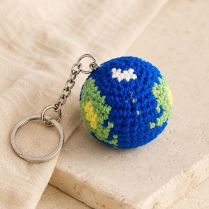 May include: A spherical crochet keychain featuring a blue, green, and yellow yarn design representing the Earth. The globe is attached to a silver-toned metal split ring by a short chain, creating a compact accessory for keys or bags.