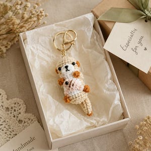 Könnte beinhalten: Ein gehäkelter Otter-Schlüsselanhänger in Beige und Weiß, der eine kleine rosa Muschel hält und an einem goldfarbenen Metallring befestigt ist. Der Anhänger liegt in einer weißen Geschenkbox mit Seidenpapier und einem Schild mit Especially for you.