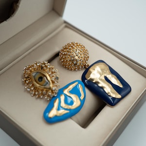 May include: A pair of statement earrings featuring a gold-tone eye motif surrounded by clear crystals, a gold-tone sphere covered in crystals, and two irregular enamel pendants in bright blue and dark navy with gold-tone abstract relief designs.
