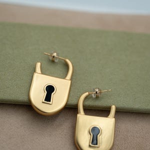 Op de afbeelding: Een paar goudkleurige oorringen in de vorm van hangsloten. Elke oorbel heeft een gladde, matte gouden afwerking met een centraal zwart sleutelgatontwerp. De oorbellen zijn voorzien van een gebogen stift en een doorzichtige kunststof sluiting.