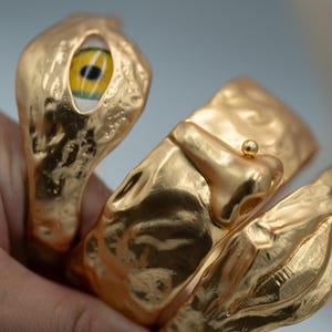 May include: A gold-toned cuff bracelet with a textured, hammered metal finish. The design features a prominent human-like eye with a yellow iris and black pupil, a sculpted nose with a small gold sphere on the tip, and a pair of lips.