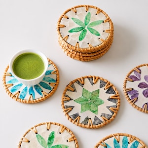May include: A collection of decorative coasters with a woven rattan border. The coasters feature various designs, including green and blue floral patterns, and a cup of green tea. The coasters are arranged on a white surface.