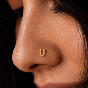 May include: A gold-toned nose stud shaped like the letter U, featuring a series of small, clear, sparkling stones embedded along the surface of the letter. The jewelry is positioned on the side of a nose, showcasing its metallic finish and design.