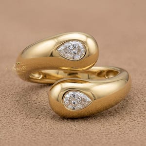 May include: A pair of gold rings with a pear-shaped diamond set in each band. The rings have a smooth, polished finish and are displayed on a neutral-toned surface. The design features a modern, elegant style.