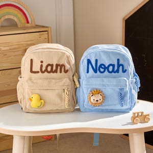 May include: Two corduroy backpacks, one beige and one light blue. The beige backpack features the name Liam in brown text and a yellow duck patch. The light blue backpack features the name Noah in blue text and a lion patch with a crown.