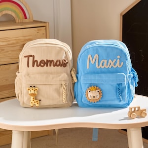 Può includere: Due zaini in velluto a coste, uno beige e uno blu, con i nomi Thomas e Maxi ricamati. Lo zaino beige ha una toppa a forma di giraffa, mentre quello blu presenta una toppa a forma di leone con corona. Entrambi hanno tiretti coordinati.