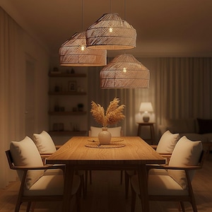 May include: Three hanging pendant lights with woven tan shades suspended above a rectangular wooden dining table. The table is surrounded by four wooden chairs with light-coloured cushions. A small vase with dried grass sits in the centre.